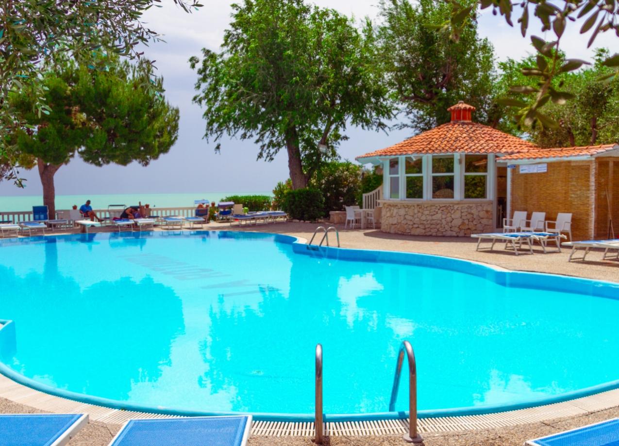 Piscina all'aperto con vista mare, circondata da lettini e alberi.