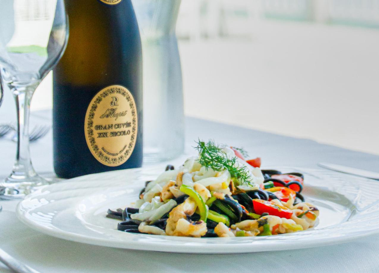 Piatto di pasta con verdure e bottiglia di vino Gran Cuvée su tavola imbandita.