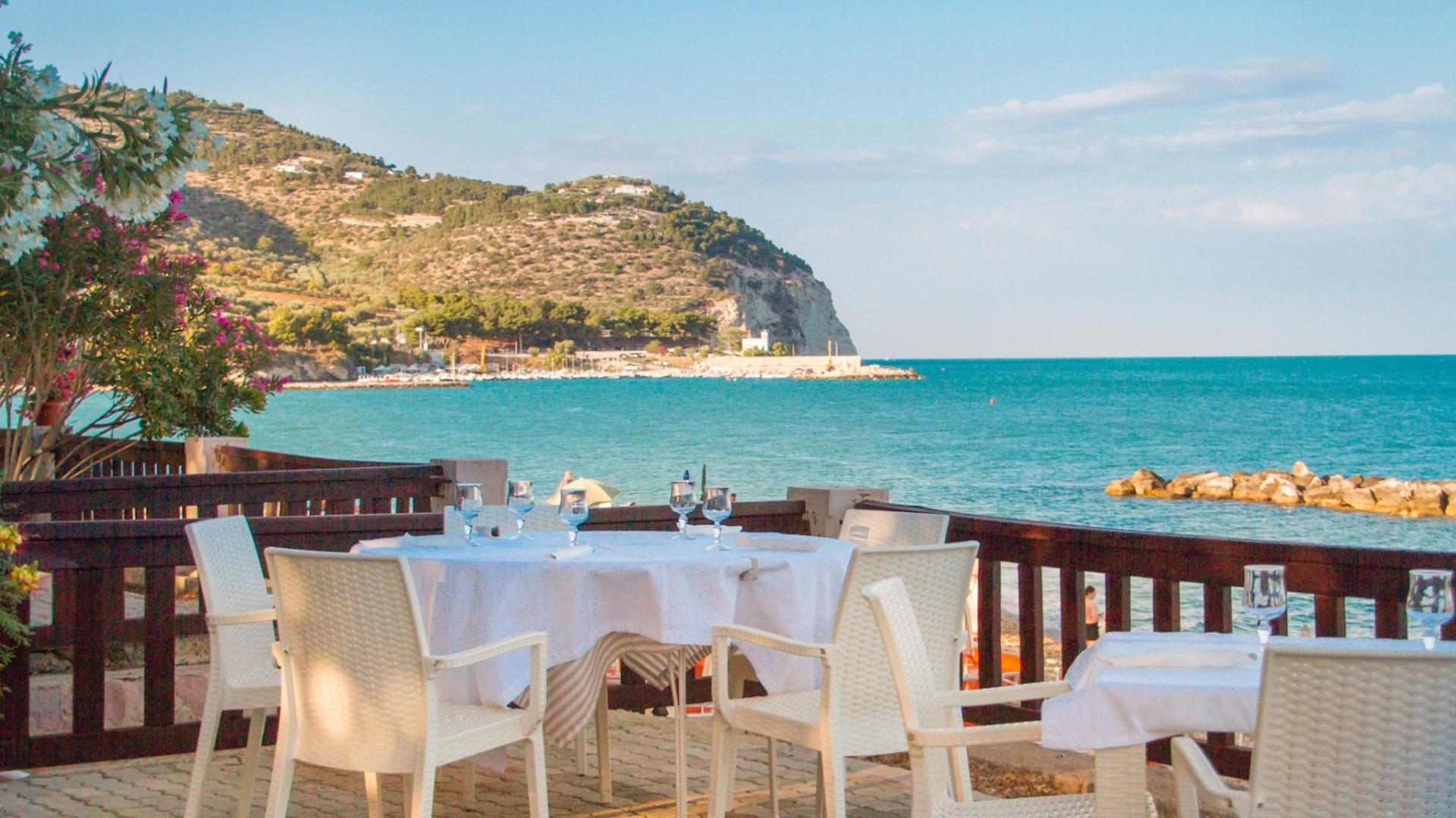 Ristorante all'aperto con vista sul mare e colline in lontananza.