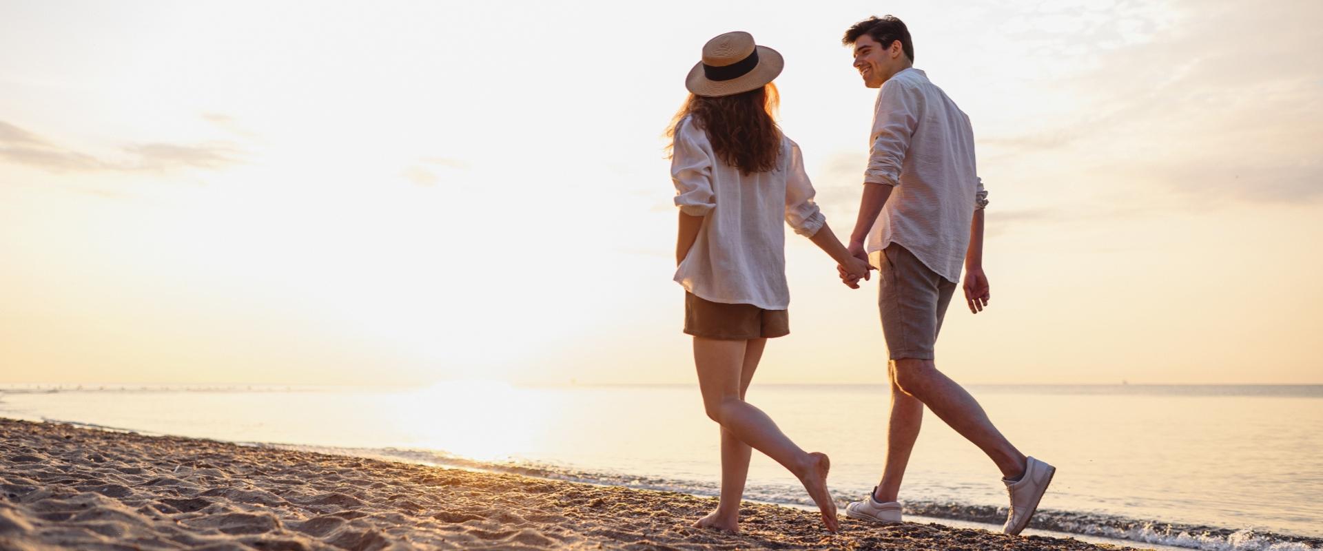 Coppia cammina sulla spiaggia al tramonto, tenendosi per mano, atmosfera romantica e serena.