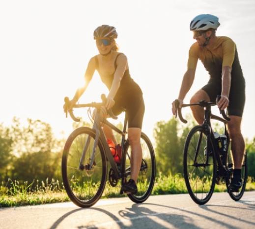 Due ciclisti pedalano insieme su una strada al tramonto, indossando caschi e abbigliamento sportivo.