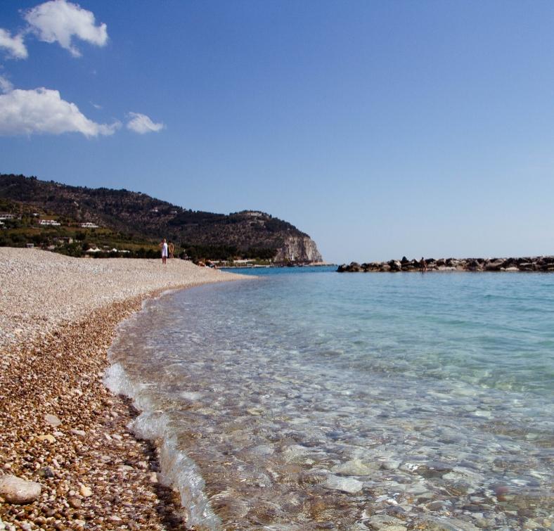 Spiaggia di ciottoli con acqua cristallina e colline sullo sfondo in una giornata soleggiata.