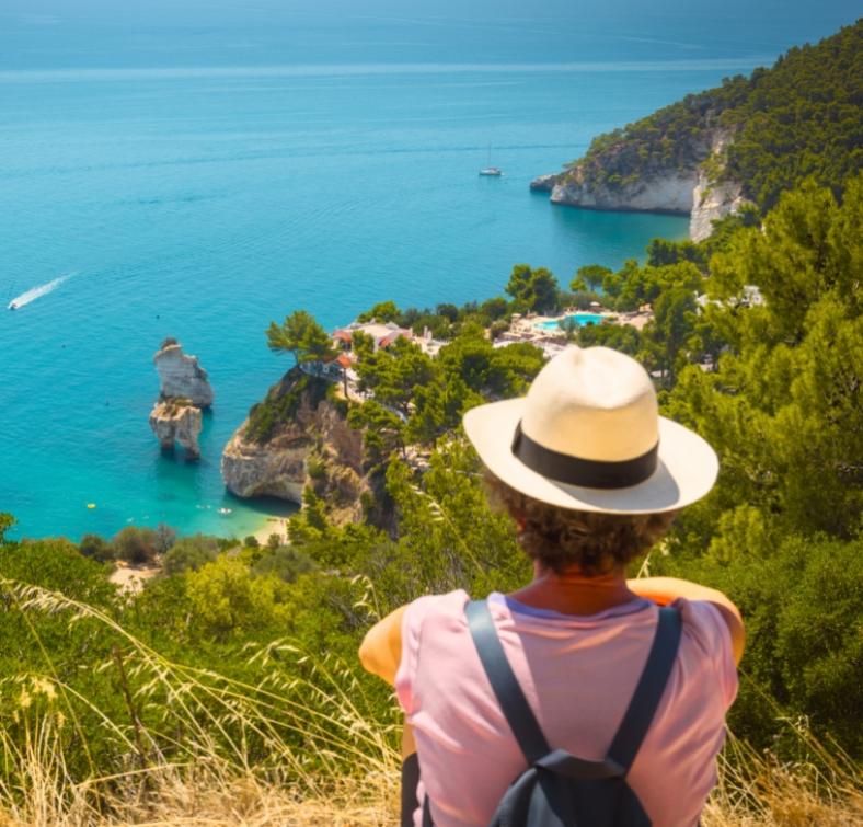 Persona con cappello osserva un paesaggio costiero con mare blu e scogli.