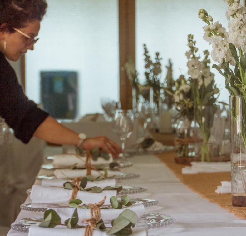 Persona apparecchia tavola elegante con fiori bianchi e tovaglioli legati con spago.