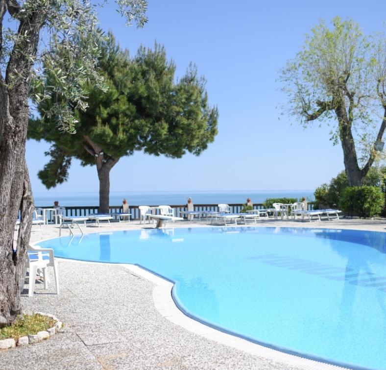 Piscina all'aperto con vista sul mare, circondata da lettini e alberi.