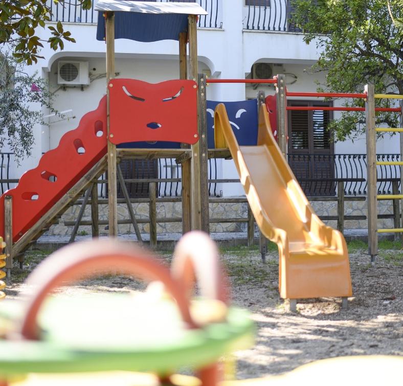 Parco giochi con scivolo arancione e struttura in legno, vicino a un edificio bianco.