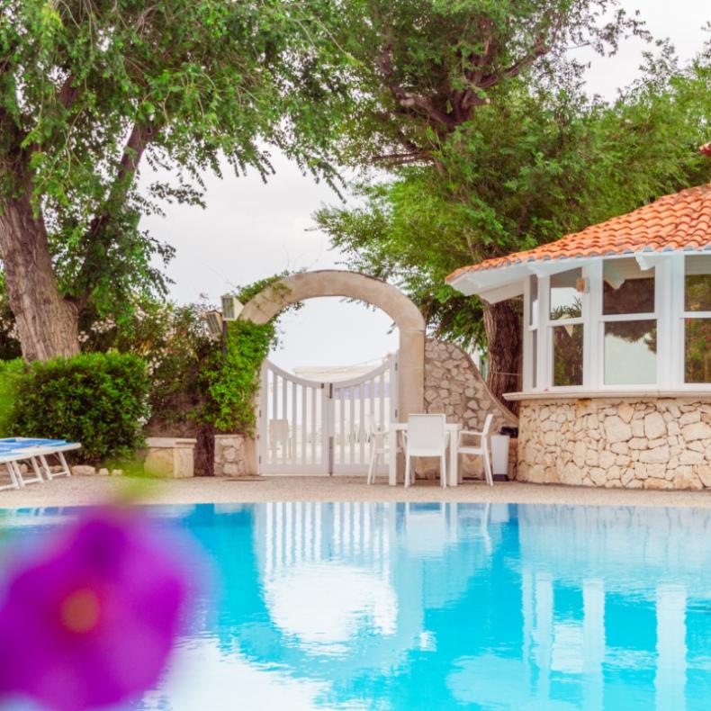 Piscina con lettini, cancello ad arco e gazebo in pietra con tetto rosso.